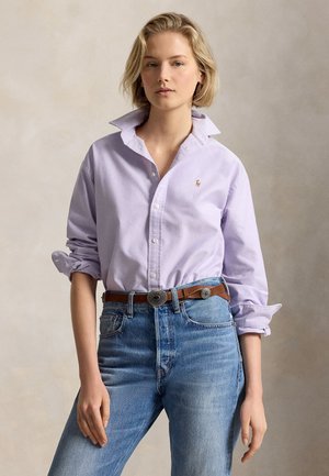 Jeune femme aux cheveux blonds courts portant une chemise boutonnée violet clair et un jean bleu avec une ceinture marron, debout devant un fond neutre.