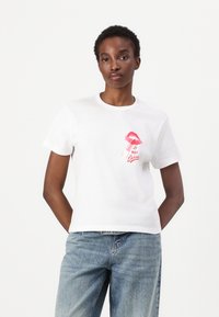 Witte katoenen t-shirt met een roze lipgrafiek en de tekst "Le Nuit Derrière", korte mouwen en een ronde hals, gecombineerd met denim.
