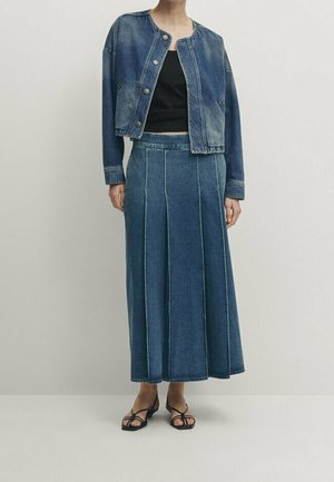 Woman wearing a cropped denim jacket, black crop top, long pleated denim skirt, and black strappy heeled sandals, standing against a plain wall.