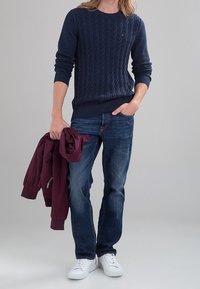 Maglione lavorato a maglia navy con motivo a treccia, abbinato a jeans in denim blu. Il modello tiene una giacca bordeaux e indossa sneakers bianche.