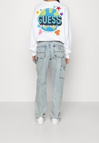 Vit sweatshirt med färggrafisk blomma och texten "GUESS ORIGINALS", ihop med ljusblå denim cargo-byxor och vita sneakers.