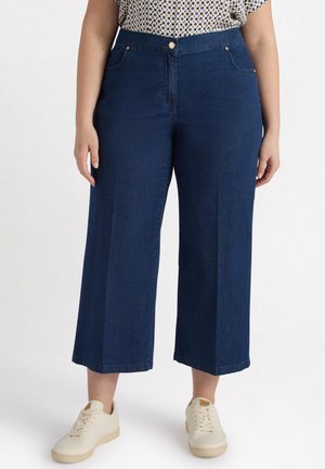 Dunkelblaue weit geschnittene Denim-Hose mit hoher Taille, zwei Fronttaschen, Knopfverschluss und rohem Saum. Kombiniert mit weißen Sneakern.