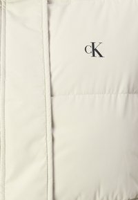 Light beige padded jacket with a smooth texture, featuring a black "cK" logo and visible stitched seams.