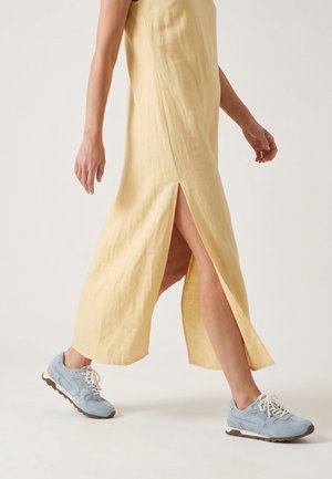 Person geht in einem gelben ärmellosen Kleid mit seitlichem Schlitz, trägt hellblaue Sneakers mit weißen Schnürsenkeln auf einem schlichten hellen Hintergrund.