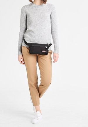 Borso nero in tessuto con chiusura a zip, indossato in vita, abbinato a un maglione grigio, pantaloni beige e sneakers bianche.