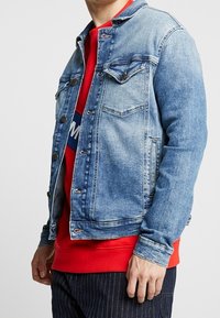 Jeansjacka i ljusblått med knäppning, två bröstfickor och en kontrasterande röd sweatshirt under, med horisontella ränder.