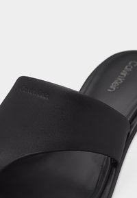 Μαύρο σαγιονάρι Calvin Klein με φαρδιά λουράκι και ανάγλυφο λογότυπο σε απλό ανοιχτό φόντο.