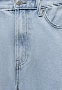 Jean en denim bleu clair en gros plan montrant la poche avant, le bouton métal, la couture de la braguette et les détails des passants de ceinture.