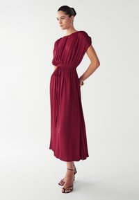 Vestido maxi burdeos con cintura fruncida, mangas cortas y aberturas laterales, hecho de una tela ligera. Combinado con sandalias de tiras negras.