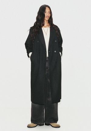 Cappotto nero in lana a doppio petto con grandi bottoni, indossato sopra un top beige e jeans in denim scuro a gamba larga, abbinato a stivali beige.