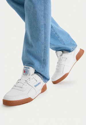 Nogi w jasnoniebieskich dżinsach noszące białe buty Reebok na gumowej podeszwie, jedna stopa lekko uniesiona na białym tle.