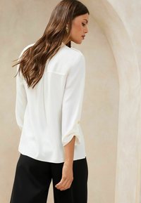 Blusa bianca con maniche a tre quarti, dettaglio dei bottoni sui polsini, texture liscia e vestibilità ampia, abbinata a pantaloni neri.