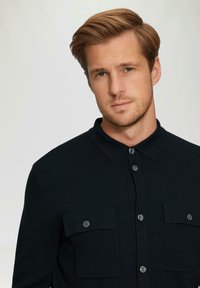 Zwarte button-up overhemd gemaakt van textuurstof; heeft twee borstzakken met knopen en een kraag.