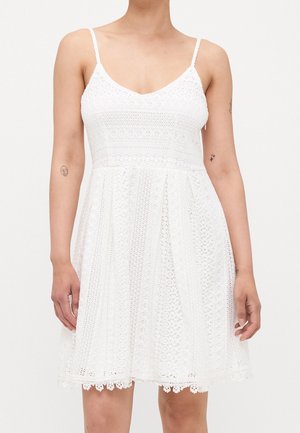 Kobieta ubrana w białą, bezrękawną koronkową sukienkę z cienkimi ramiączkami i rozkloszowanym dołem, ukazująca tatuaże na obu przedramionach.