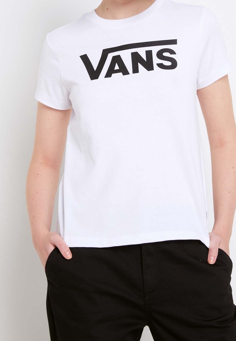 Personne portant un t-shirt Vans blanc avec un logo noir, les mains dans les poches, associé à un pantalon noir, sur un fond uni clair.