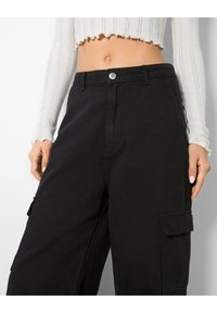 Mustat cargo-housut, jotka on valmistettu paksusta kankaasta ja joissa on väljä istuvuus. Edessä napitus ja sivutaskut. Teksturoitu vyötäröyksityiskohta näkyvissä.