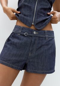 Denimshorts i mörkblått med kontrasterande gula sömmar. Har knäppning, bälteshällor och en slät yta.