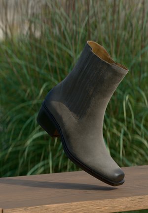 Gray suede ankle boot with block heel floating above a wooden bench, tall grass blurred in the background.
