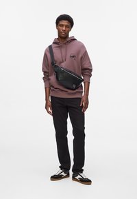 Geantă de umăr neagră, cu suprafață texturată, purtată pe umăr. Model îmbrăcat cu hanorac violet, blugi negri și adidași negri cu portocaliu. Elemente de design simple.