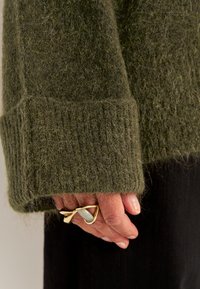 Olijfgroene wollen trui met geribbelde manchetten en een fuzzy textuur. Hand die een gouden ring vasthoudt met een stenen accent. Zwarte stof zichtbaar eronder.