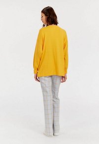 Pull en maille jaune à coupe oversize, associé à un pantalon à carreaux bleu clair et beige. Mannequin vu de dos, portant des baskets blanches.