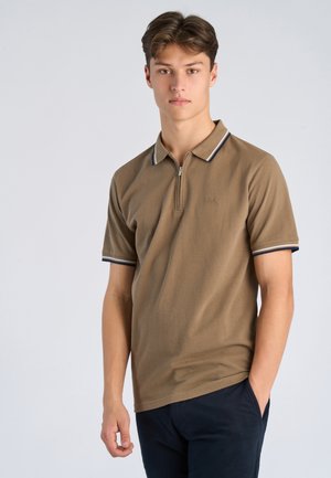 Młody mężczyzna ubrany w brązowe polo z krótkim rękawem i suwakiem, granatowe spodnie, stoi z jedną ręką w kieszeni na jasnoszarym tle.