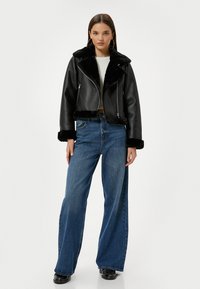 Giacca nera in pelle corta con colletto e polsini in pelliccia sintetica, abbinata a jeans a gamba larga blu. Il modello si trova su uno sfondo neutro.
