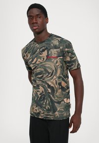 Joven vestido con una camiseta de patrón abstracto en verde y beige con el texto rojo "JUST CAVALLI", combinada con pantalones negros, frente a un fondo liso.