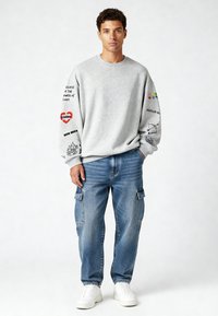 Jauns vīrietis stāv, ģērbies pelēkā džemperī ar apdrukātu tekstu un grafiku uz piedurknēm, zilās kravas biksēs un baltās kedās.