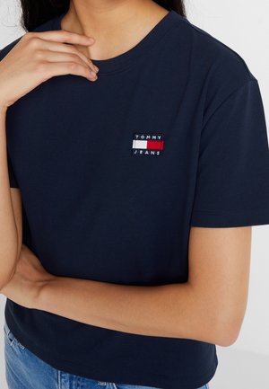 Frau trägt ein marineblaues Tommy Jeans T-Shirt mit kurzer Ärmeln und Logo-Patch auf der Brust, kombiniert mit hellblauen Denim-Jeans.