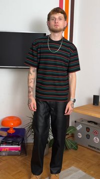 Camisa de manga curta listrada em preto e vermelho com mangas largas, combinada com calças de couro pretas largas. Apresenta tatuagens visíveis nos braços.