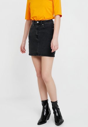 Femme portant une jupe courte en jean noire, un t-shirt orange vif à manches courtes et des bottines noires brillantes, debout sur un fond blanc.