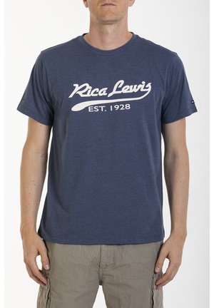 Homme portant un t-shirt bleu marine "Rica Lewis EST. 1928" et un short cargo beige, debout avec les bras détendus le long du corps.