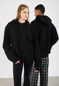 Fekete, bolyhos fleece zipzáros kapucnis pulóver, elülső zsebbel és állítható kapucnival; fekete, testhezálló nadrággal viselve, háttérben kockás nadrágokkal.