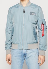 Light blue bomber jacket with a front zipper, ribbed cuffs and hem, side pockets, and a red fabric tag on the sleeve.