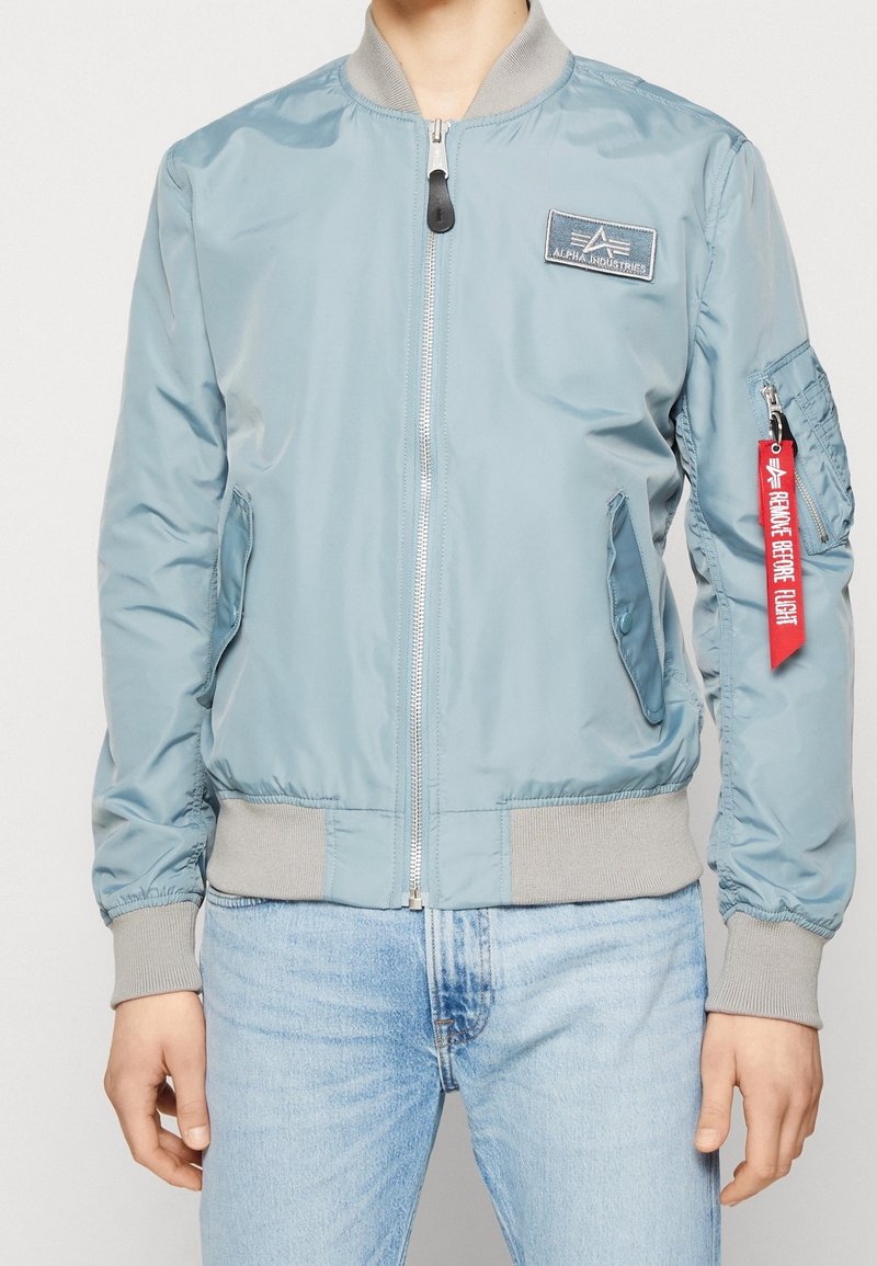 Light blue bomber jacket with a front zipper, ribbed cuffs and hem, side pockets, and a red fabric tag on the sleeve.