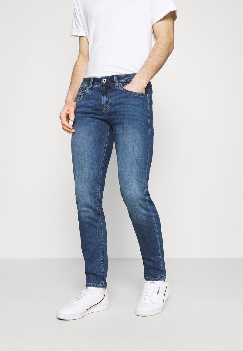 Slim-fit blue denim jeans, cotton fabric, faded finish, five-pocket design, zipper fly, worn with a white T-shirt and white sneakers.