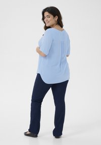 Blouse bleu clair à manches courtes, coupe ample, légère texture, ourlet arrondi. Associée à un pantalon bleu foncé et des chaussures noires.