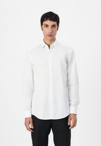 Cămașă albă cu mâneci lungi, din bumbac, având un guler clasic și un sistem de închidere cu nasturi; asortată cu pantaloni negri. Textură netedă și croială.