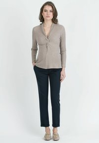 Frau, die vor einem weißen Hintergrund steht und einen beigen, gedrehten, gerippten Pullover, schwarze Hosen und Mokassins aus Schlangenleder trägt.