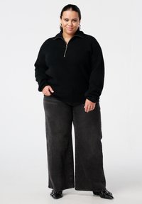 Zwarte gebreide trui met een kwart-rits ontwerp, gecombineerd met wijd uitlopende donkergrijze jeans en zwarte schoenen met puntige neus. Simpele zilveren sieraden als accenten.