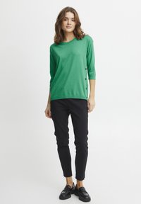 Zeleni pleteni top z okroglo izrezom, s tričetrtinskimi rokavi in stranskimi gumbnimi detajli, v kombinaciji s črnimi ožjimi hlačami in črnimi loaferji.