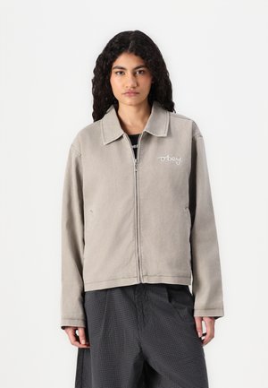 Sivi zip-up sako s oštrim ovratom i vezom "obey" logotipa na lijevom prsnom dijelu. Ima dugačke rukave i opušteni kroj.