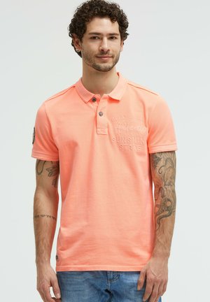 Man with curly hair and tattoos wearing a coral polo shirt and blue jeans, standing against a plain light background.