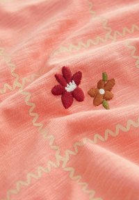 Korallfärgad tyg med två broderade blommor: en röd med en vit mitt och en brun med en vit mitt, accentuerad av ett zigzag-sticks mönster.