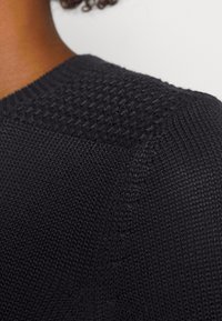 Närbild av en persons axel som bär en svart strukturerad stickad tröja med mönstrade sömdetaljer på övre delen av ryggen och ärmen.