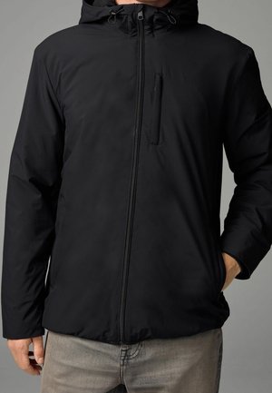 Homme portant une veste à capuche noire avec une fermeture éclair à l'avant et une poche poitrine, la main dans la poche latérale, sur un fond gris.