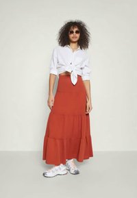 Μακρύ κόκκινο φουστάνι με επίπεδη σιλουέτα, σε συνδυασμό με λευκό πουκάμισο δεμένο και χοντρές αθλητικές παπούτσια, που αποπνέει μια αθλητική αισθητική.