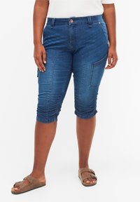 Pantalones capri de mezclilla con un diseño ajustado, color azul, bolsillos laterales y detalle recogido en los puños. Se llevan con sandalias marrones.