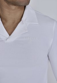 Fehér, hosszú ujjú pólóing, galléros nyakkal. Finom hímzett logóval a mellrészen és sima, testhezálló textúrával rendelkezik.